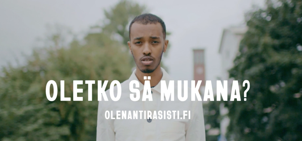 Kuvassa tummaihoinen miesoletettu kesäisessä kaupunkimaisemassa. Kuvassa kysymys "Oletko sä mukana?" sekä kampanjan verkkosivujen osoite olenantirasisti.fi.